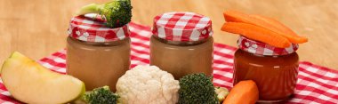 Panoramic shot of jars of baby food with fresh vegetables and apple on tablecloth on wooden surface