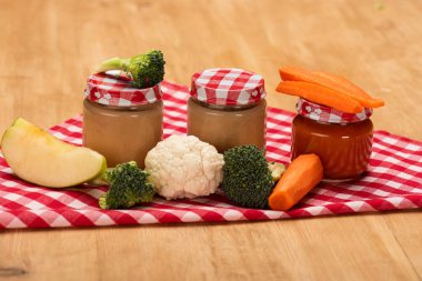 Jars of vegetable baby nutrition with fresh vegetables and apple on napkin on wooden surface