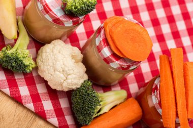 High angle view of jars of vegetable baby food with fresh vegetables and apple on tablecloth on wooden surface