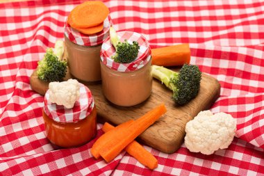 Baby nutrition in jars with carrots, broccoli and cauliflower on cutting board on checkered tablecloth