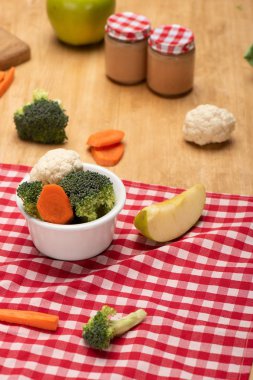 Selective focus of fresh vegetables and apple on tablecloth with jars of baby nutrition on wooden surface