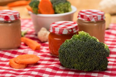 Close up view of fresh broccoli, carrot and cauliflower with jars of baby food on tablecloth on wooden surface