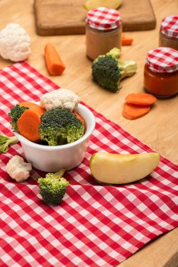 Organic vegetables and apple on napkin near jars of baby food on wooden background