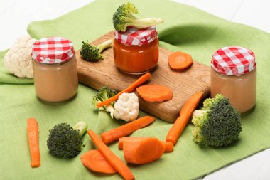 Jars of baby food with ripe carrots, broccoli and cauliflower on napkin on white wooden background