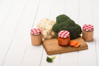 Jars of baby food with broccoli, cauliflower and carrot slices on cutting board on white wooden surface