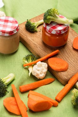 High angle view of carrot, cauliflower and broccoli with jars of baby food on cutting board on tablecloth