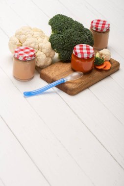 Baby nutrition in jars with ripe vegetables and spoon on cutting board on white wooden surface
