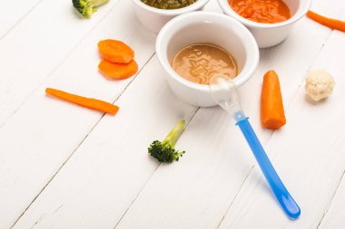 Bowls with baby food with pieces of vegetables and spoon on white wooden background