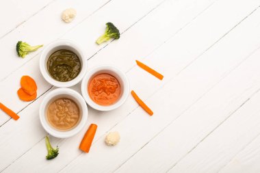 Top view of bowls with baby food with vegetables on white wooden background