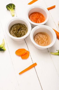 High angle view of pieces of carrots and broccoli near bowls of baby food on white wooden background