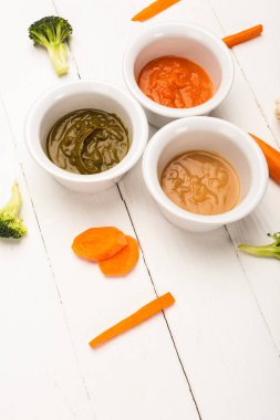 High angle view of pieces of carrots and broccoli near bowls of baby food on white wooden background clipart