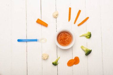 Top view of vegetable baby nutrition in bowl with pieces of fresh vegetables and spoon on white wooden surface