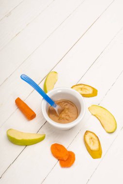 High angle view of baby food in bowl with spoon and sliced fruits and carrot on white wooden background