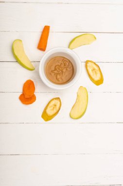 Top view of baby food in bowl with sliced fruits and carrot on white wooden surface
