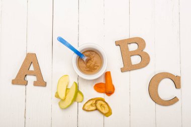 Top view of baby food with spoon and ingredients near wooden letters on white wooden background