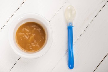 Top view of bowl with baby food and spoon on white wooden background