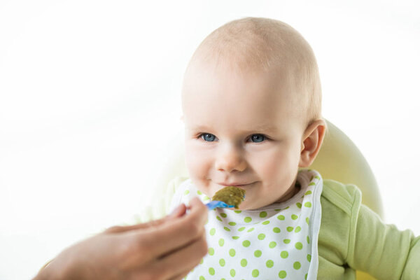 Selective focus of infant looking at mother with spoon while eating on feeding chair isolated on white