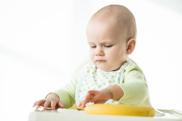 Selective focus of baby sitting on feeding chair with banana on table on white background
