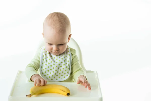 Infant touching ripe banana while sitting on feeding chair on white background