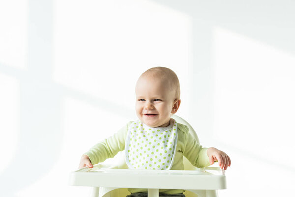 Cheerful baby sitting on feeding chair and smiling away on white background