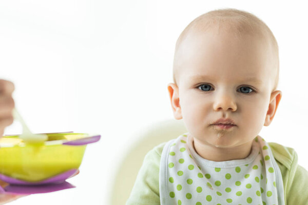 Selective focus of infant on feeding chair looking at camera near mother with bowl of puree isolated on white