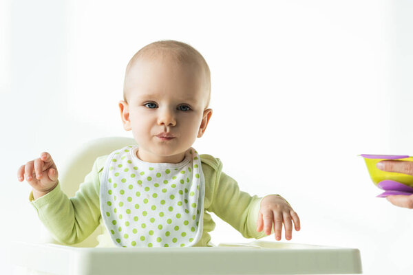 Adorable baby looking at camera while sitting on feeding chair near mother with bowl of baby nutrition on white background