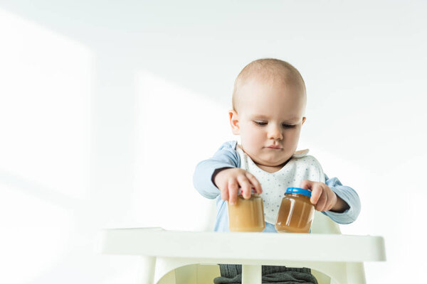 Cute baby holding jars of baby food on table of feeding chair on white background