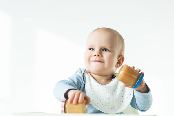 Selective focus of baby holding jars of fruit baby nutrition and smiling away while sitting on feeding chair on white background