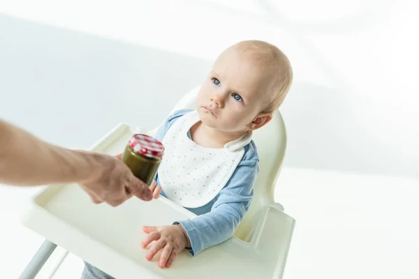 High angle view of mother giving jar of baby nutrition to baby son on high char on white background