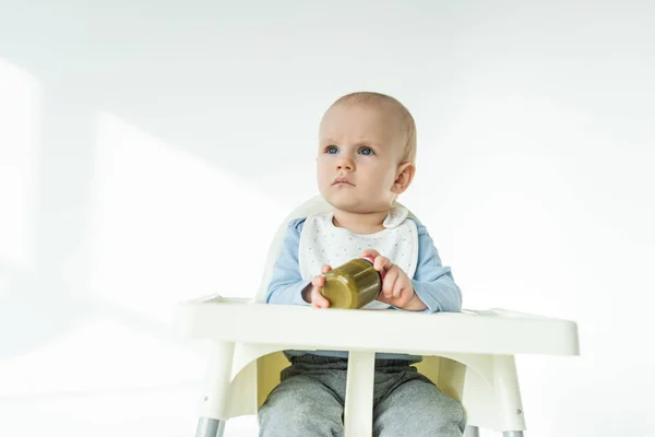 Cute baby boy holding jar of vegetable puree on feeding chair on white background