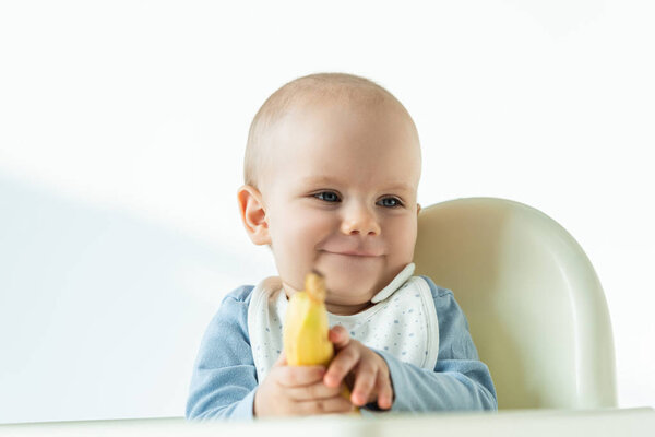 Selective focus of smiling baby holding banana while sitting on feeding chair on white background