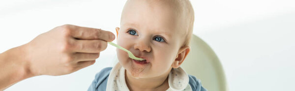 Panoramic shot of mother with spoon feeding cute baby boy on feeding chair on white background