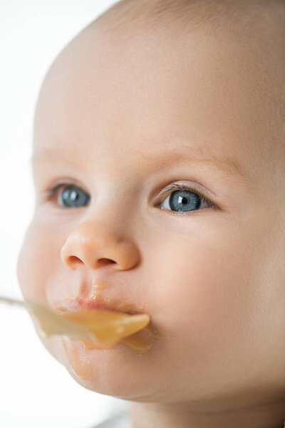 Selective focus of cute baby looking away during feeding with fruit puree isolated on white 