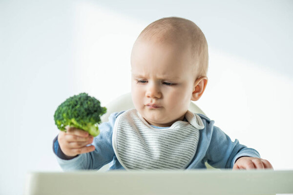 Selective focus of baby holding piece of broccoli while sitting on feeding chair on white background
