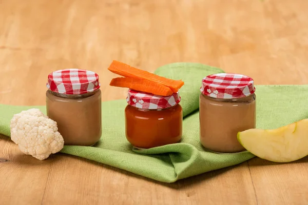 Pieces of apple, cauliflower and carrot near jars of baby food on napkin on wooden surface