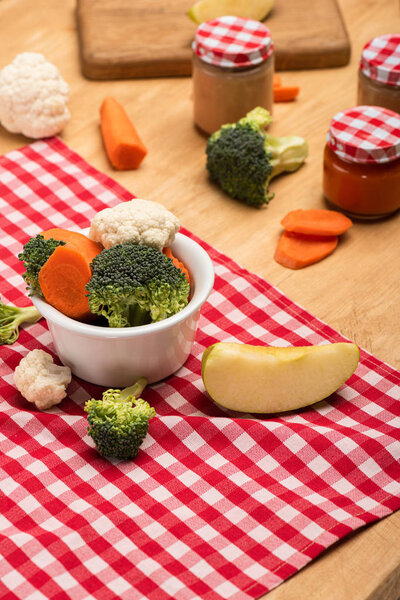 Organic vegetables and apple on napkin near jars of baby food on wooden background
