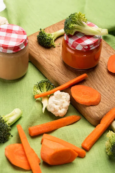 High angle view of carrot, cauliflower and broccoli with jars of baby food on cutting board on tablecloth