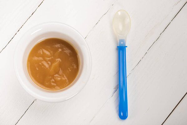 Top view of bowl with baby food and spoon on white wooden background