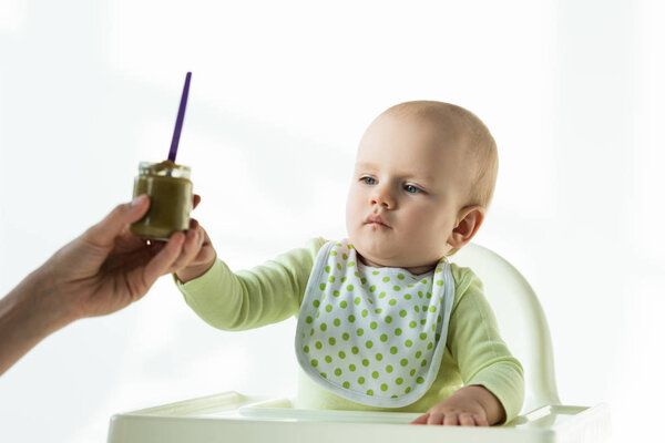 Selective focus of mother giving jar of baby nutrition to infant on feeding chair on white background