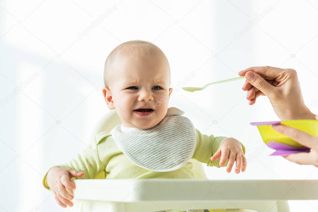 Upset baby on feeding chair looking at mother with bowl of baby nutrition on white background