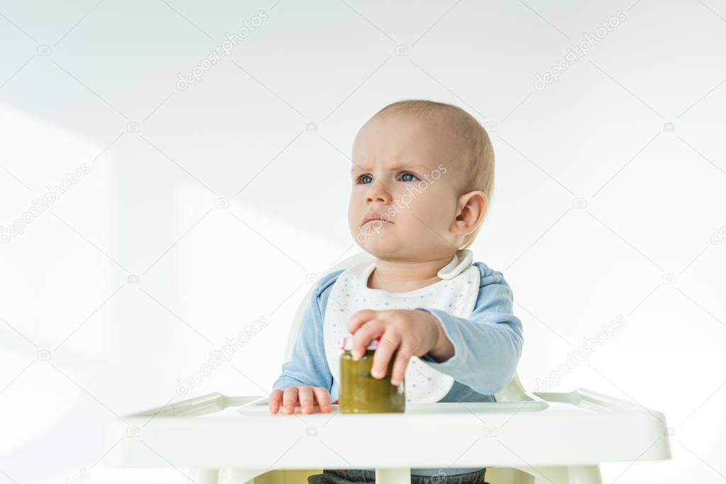 Cute baby boy holding jar of vegetable puree and looking away on feeding chair on white background