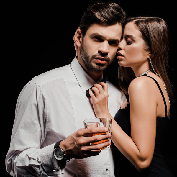 attractive woman touching bow tie of handsome man with glass of whiskey isolated on black 