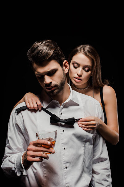 attractive woman holding bow tie near man with glass of whiskey isolated on black 