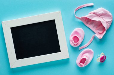 Ahşap çerçevedeki boş karatahta, pembe bebek kıyafetleri ve mavi arka planda emzik, anneler günü konsepti. 