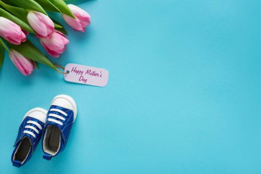 Üzerinde mutlu anneler günü yazan lalelerin üst görüntüsü mavi arka plandaki bebek paçalarının yanında yazılı kağıt etiketler.