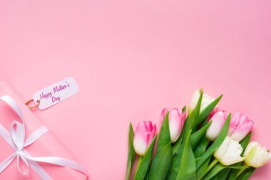 Hediyenin üst görüntüsü mutlu anneler günü kağıda yazılmış pembe arka plandaki lalelerin yanında.