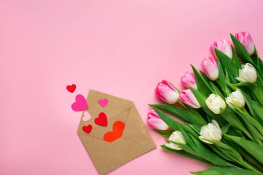 Üzerinde kağıt kalpler olan pembe yüzey üzerindeki lale demetinin yanındaki zarfın üst görüntüsü