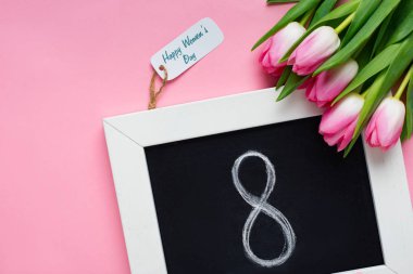 Üzerinde 8 numara olan yazı tahtasının üst görüntüsü, mutlu kadınlar günü harfleri olan kağıt etiketi ve pembe yüzey üzerinde laleler.