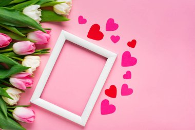 Kağıt kalplerle beyaz çerçevenin üst görüntüsü ve pembe yüzeyde bir buket lale.