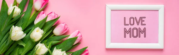 Top view of love mom lettering in frame and bouquet of tulips on pink surface, panoramic shot